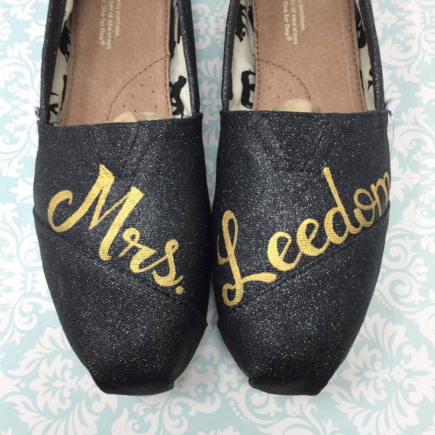 black glitter wedding toms with Mrs painted in gold. light blue paisley patterned background
