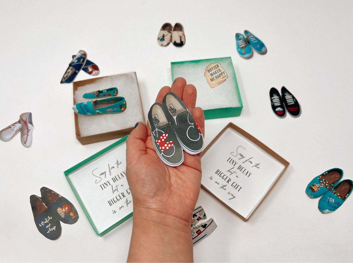 a hand holding a pair of mini shoes in its palm. There are more mini shoes & gift boxes in the back with ButterMakesMeHappy logo